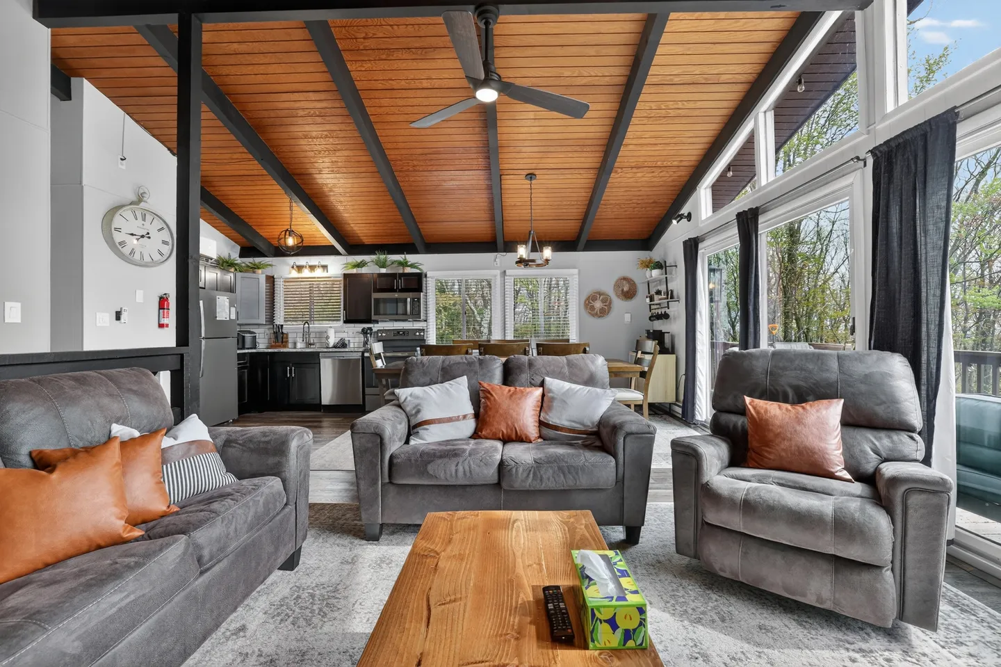Open-concept living room with gray upholstered seating, wood-plank ceiling, and a view into the kitchen and dining area.