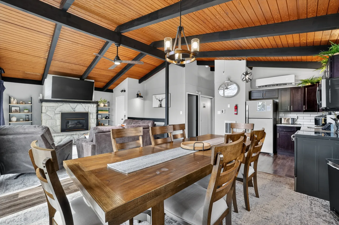 Open-concept living space with a wooden dining table in the foreground, adjacent kitchen, and living room with a stone fireplace under exposed wood-beam ceilings.