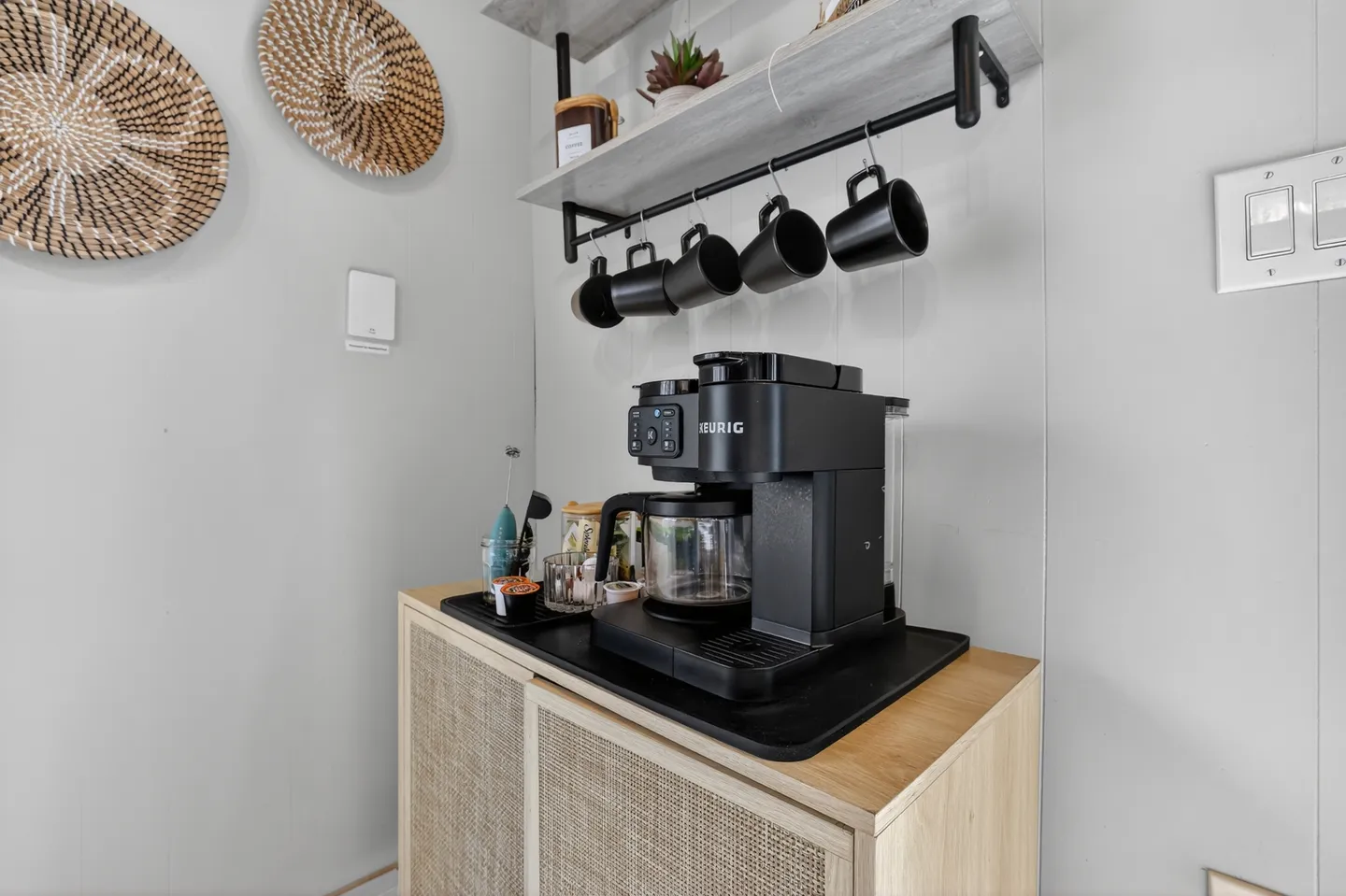 Dedicated coffee station featuring a black coffee maker on a light wood cabinet with hanging mugs above.