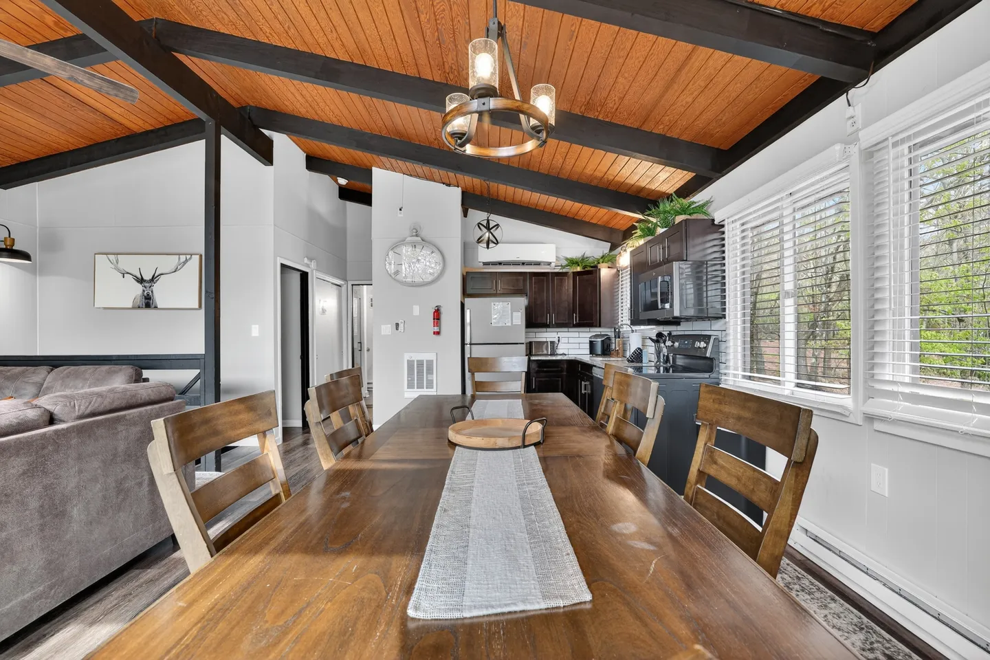 Dining area with a long wooden table and chairs beneath a wood-paneled vaulted ceiling with exposed beams, opening toward the kitchen and living room.
