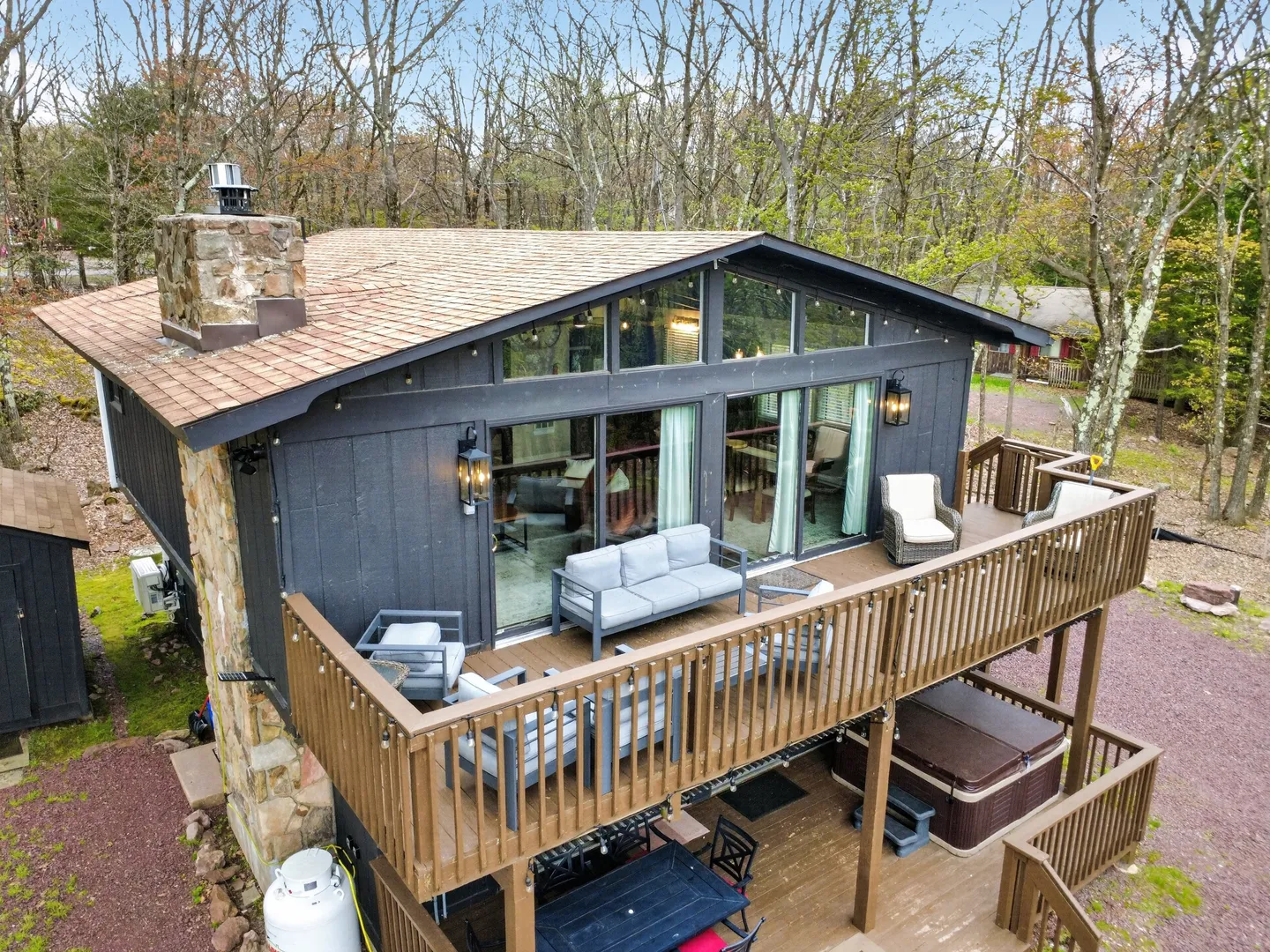 Elevated view of a dark wood cabin exterior featuring a multi-level deck with outdoor seating, a hot tub below, a stone chimney, and large upper windows.