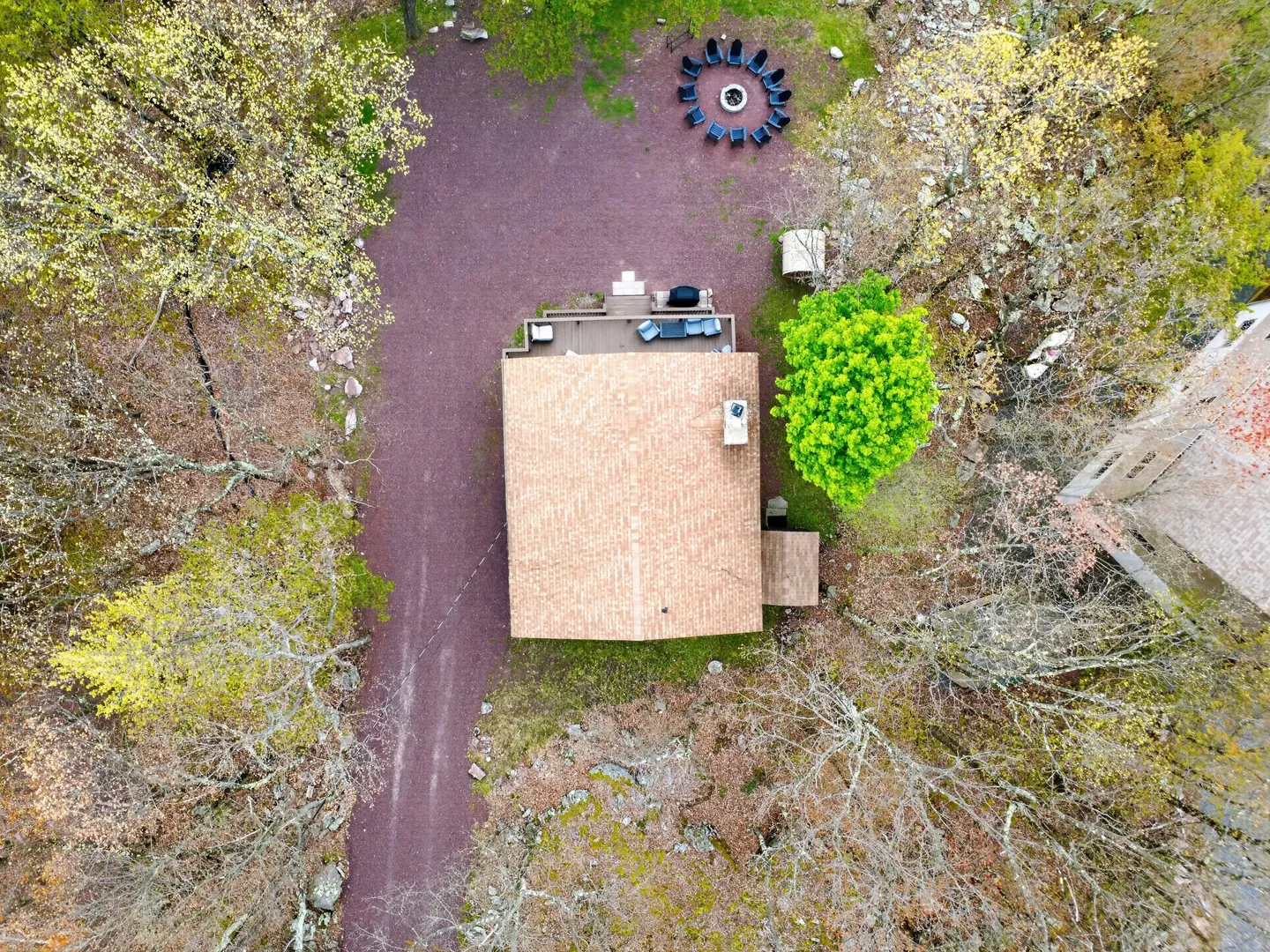 Bird's-eye aerial view of a cabin with a brown shingled roof, gravel driveway, surrounding woods, and an outdoor fire pit with a ring of Adirondack chairs.