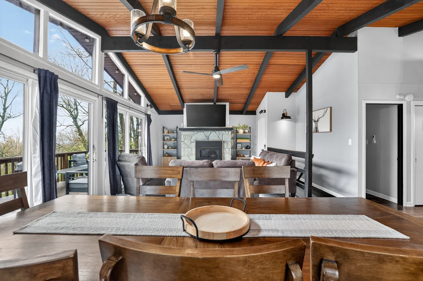 Open-concept living and dining area with vaulted wood ceiling, dark beams, stone fireplace, and floor-to-ceiling windows.