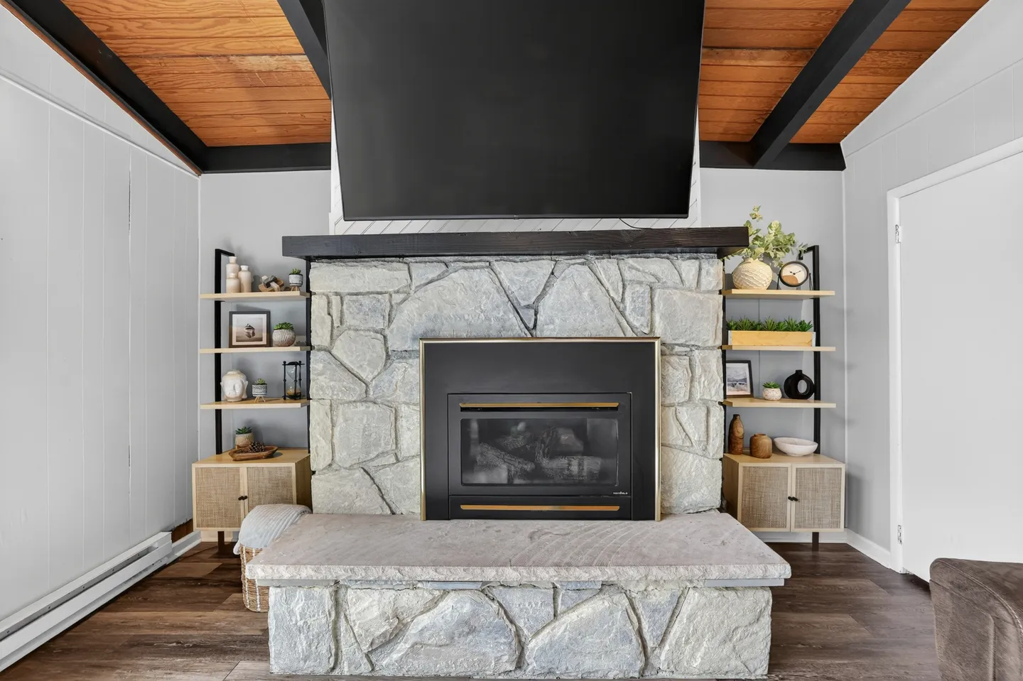 A stone fireplace with a raised hearth, mounted television above the dark mantel, and open shelving units on both sides beneath an exposed wood beam ceiling.