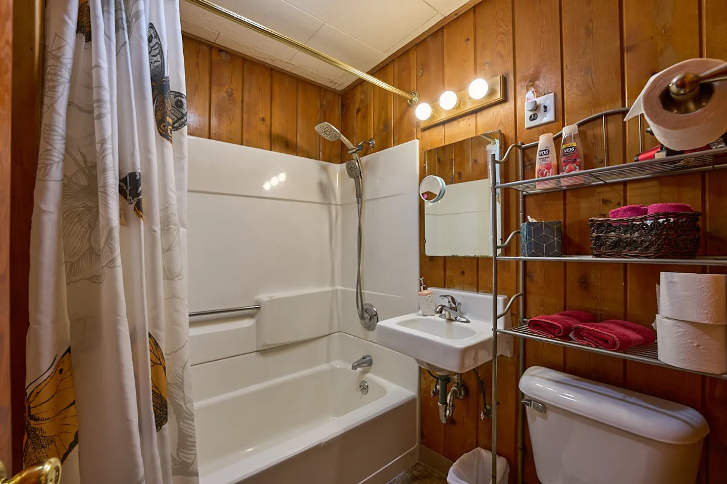 Bathroom with wood-paneled walls, white bathtub and shower surround, wall-mounted sink, and toilet with an over-the-tank metal shelving unit.