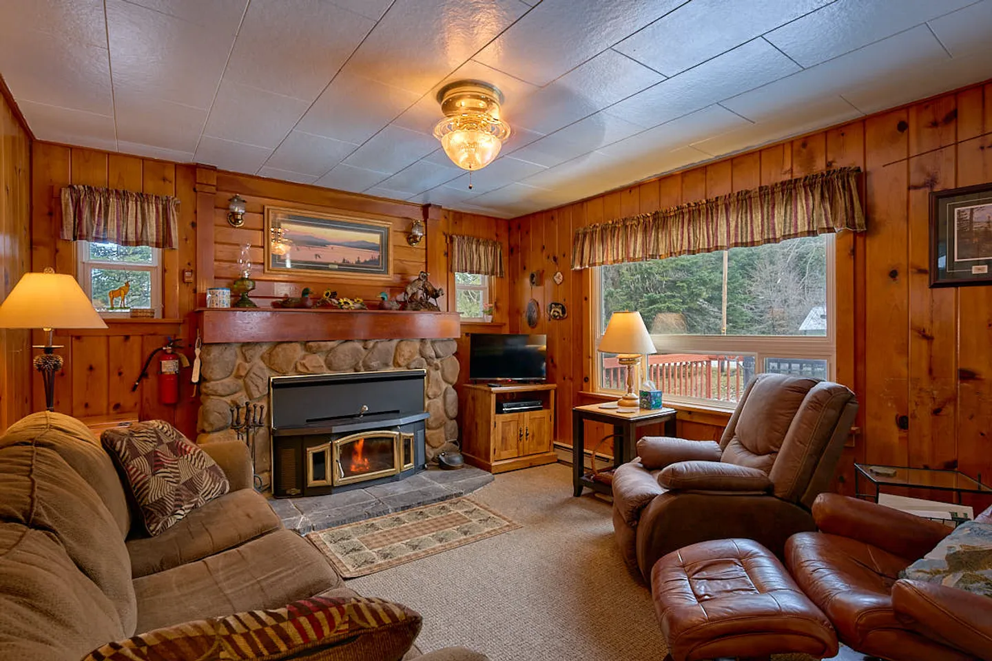 Living room with wood-paneled walls, a stone fireplace with a wood-burning insert, upholstered armchairs, and a sofa.