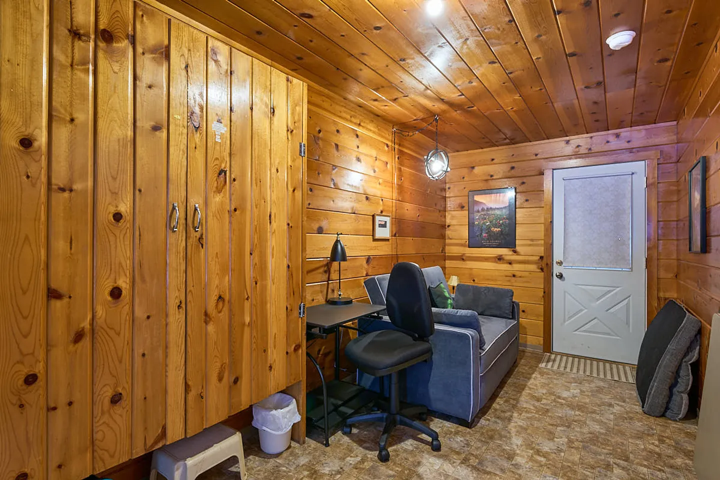 Wood-paneled room featuring a desk with an office chair, a blue armchair, a large wooden cabinet, and an exterior door.