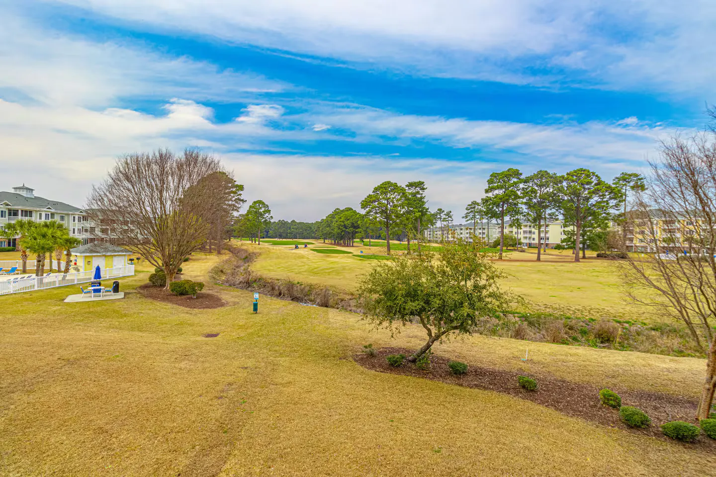 Elevated view overlooking a golf course, manicured lawns, trees, nearby residential buildings, and a fenced community pool area under a blue sky.