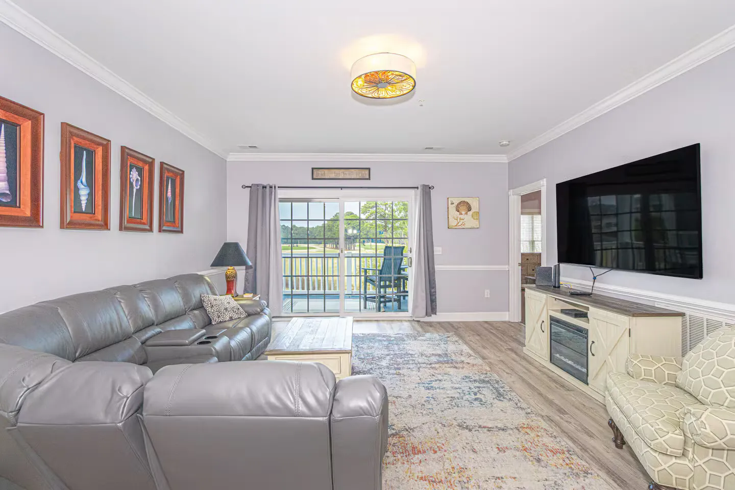 Living room with a large gray sectional sofa, TV mounted above a console with an electric fireplace, patterned armchair, and sliding glass doors leading to a balcony.