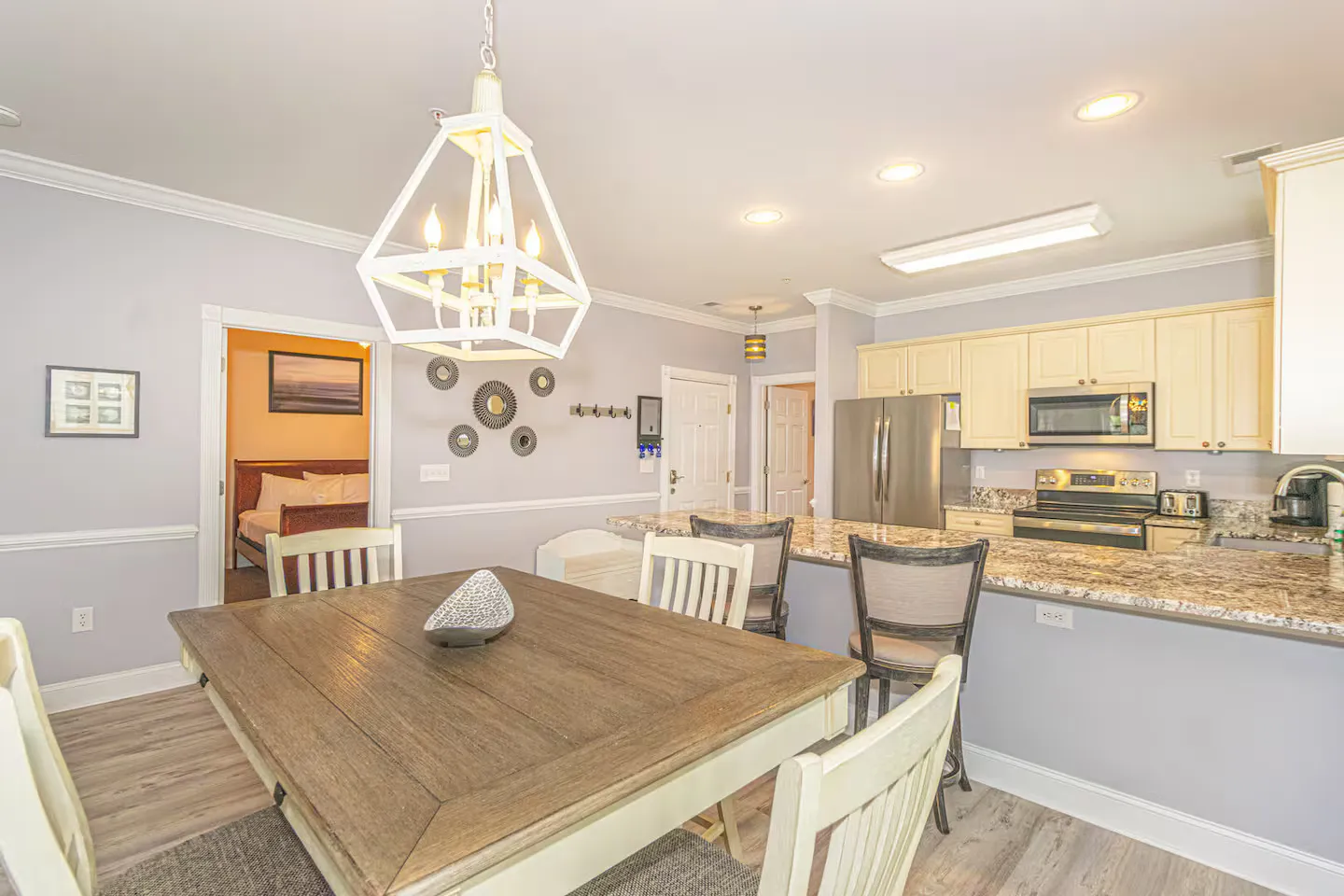 Dining area with a wood-top table and geometric chandelier, adjacent to a kitchen with granite counters and light cabinetry.