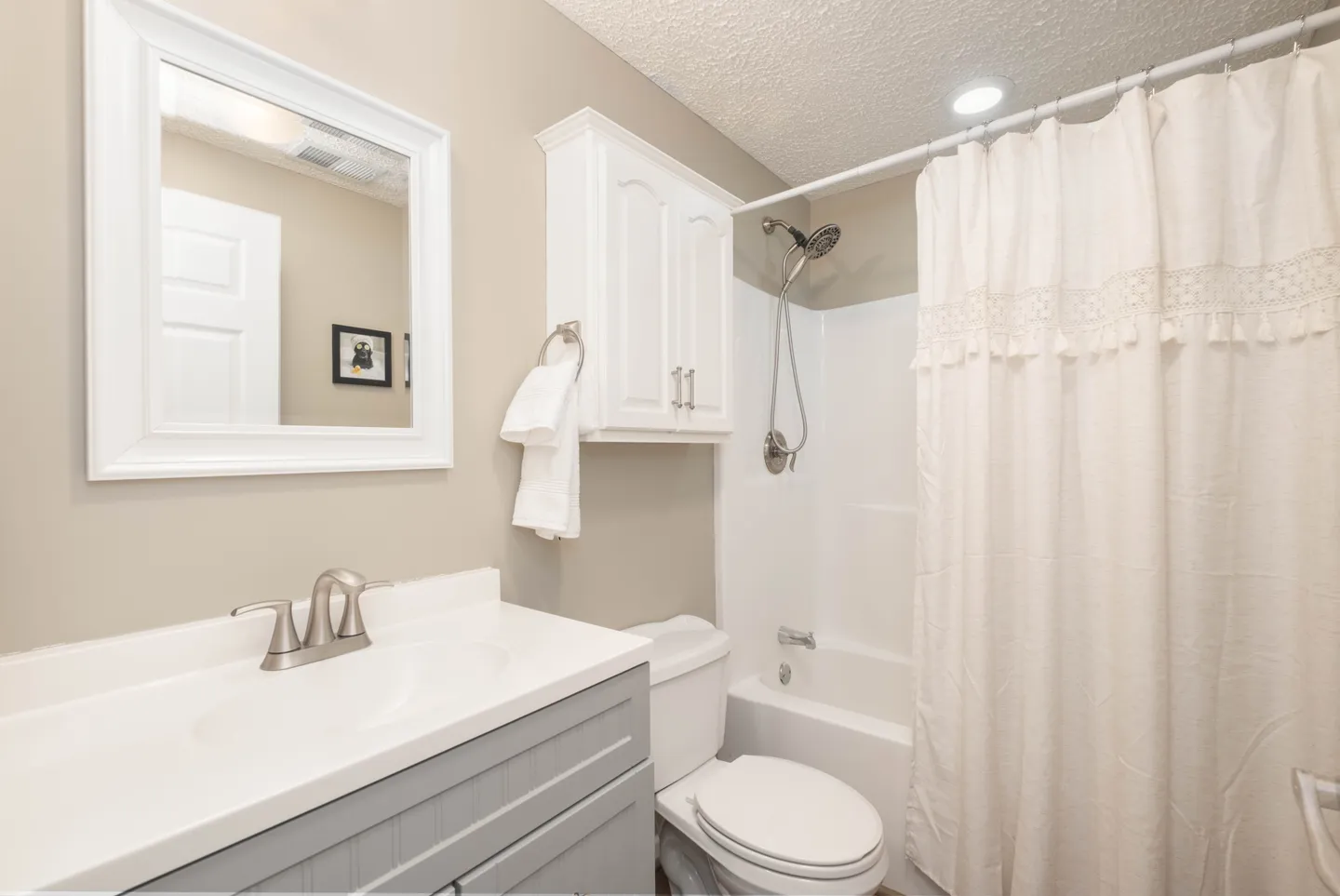 Bathroom with a grey vanity, white mirror, toilet, wall cabinet, and shower-tub combination with a white curtain.