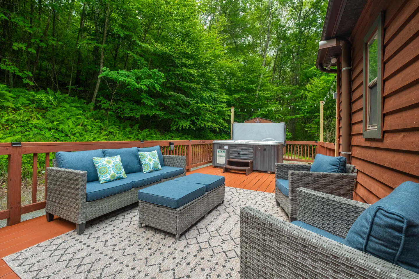 Outdoor wooden deck featuring a wicker seating set with blue cushions, an outdoor rug, and a covered hot tub surrounded by dense green forest.