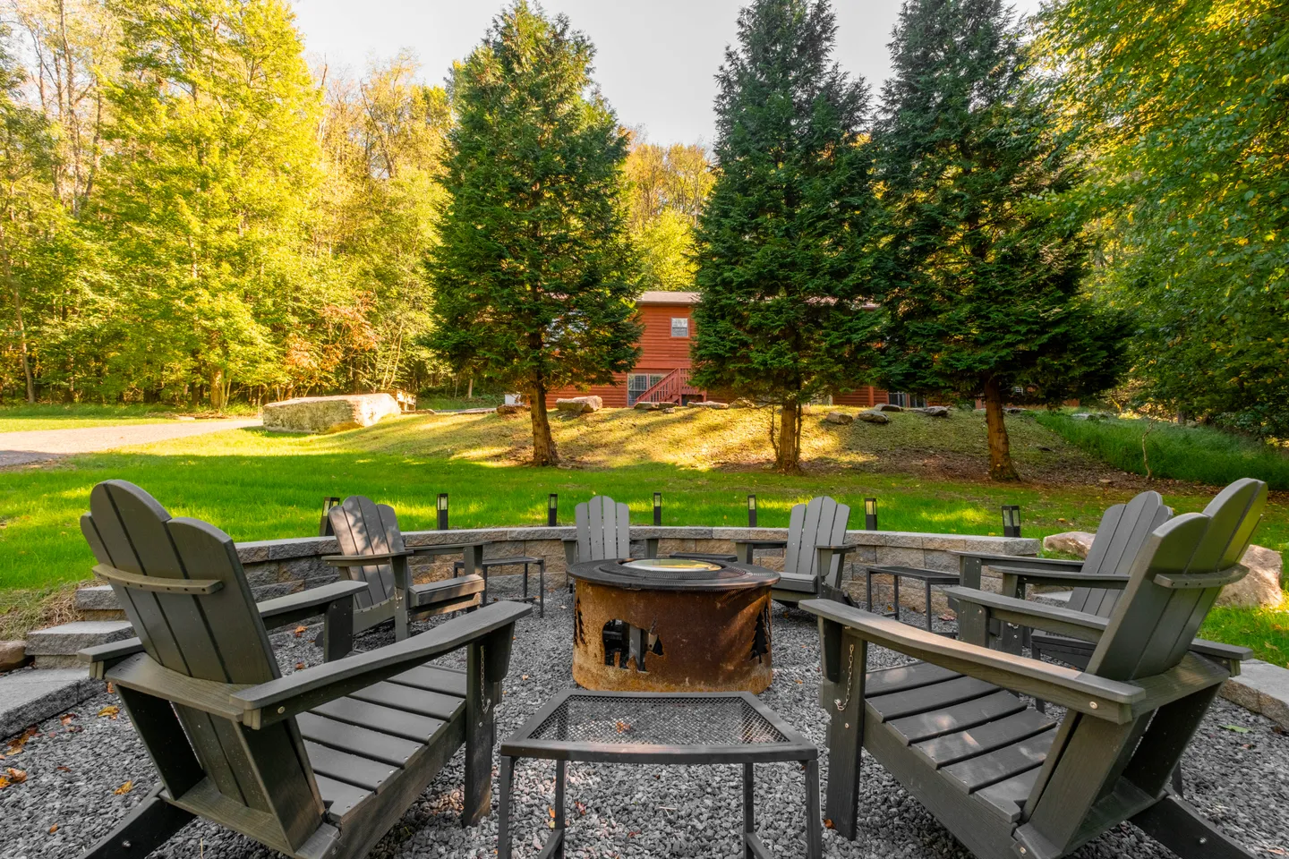 Outdoor sunken fire pit area with gravel ground, Adirondack chairs, and views of a grassy lawn and trees.