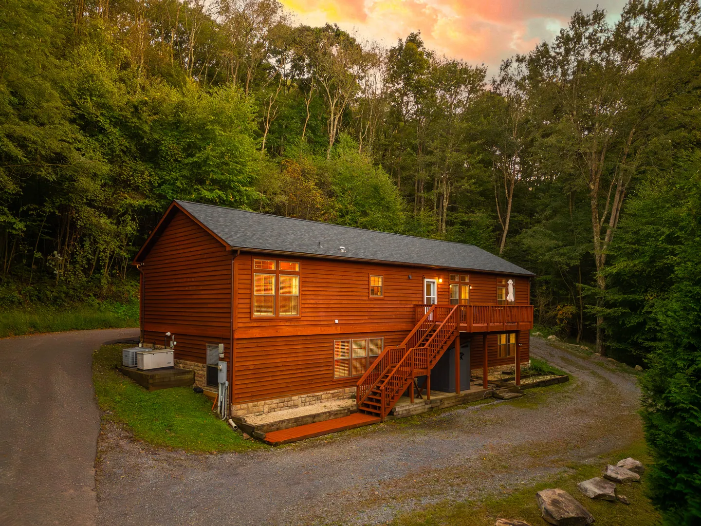 Exterior of a two-story wooden cabin with an elevated wooden deck surrounded by dense forest at sunset.