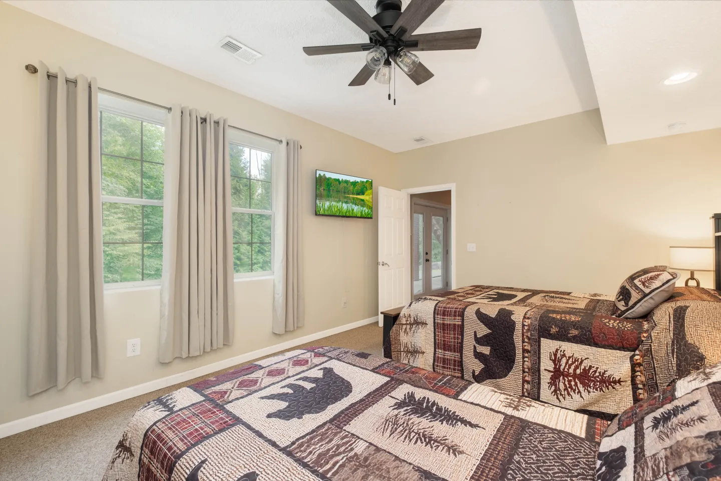 Bedroom with two beds covered in rustic bear-patterned quilts, carpeted floors, two windows with grey curtains, and a ceiling fan.
