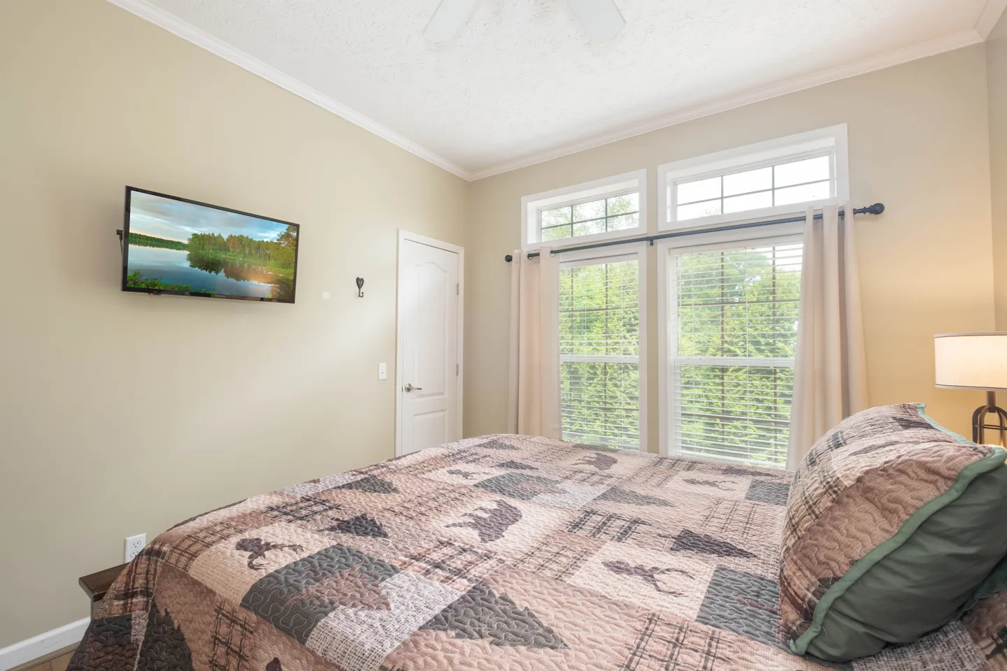 Bedroom featuring a bed with a nature-themed quilt, a wall-mounted TV displaying a lake image, and windows with white blinds overlooking trees.