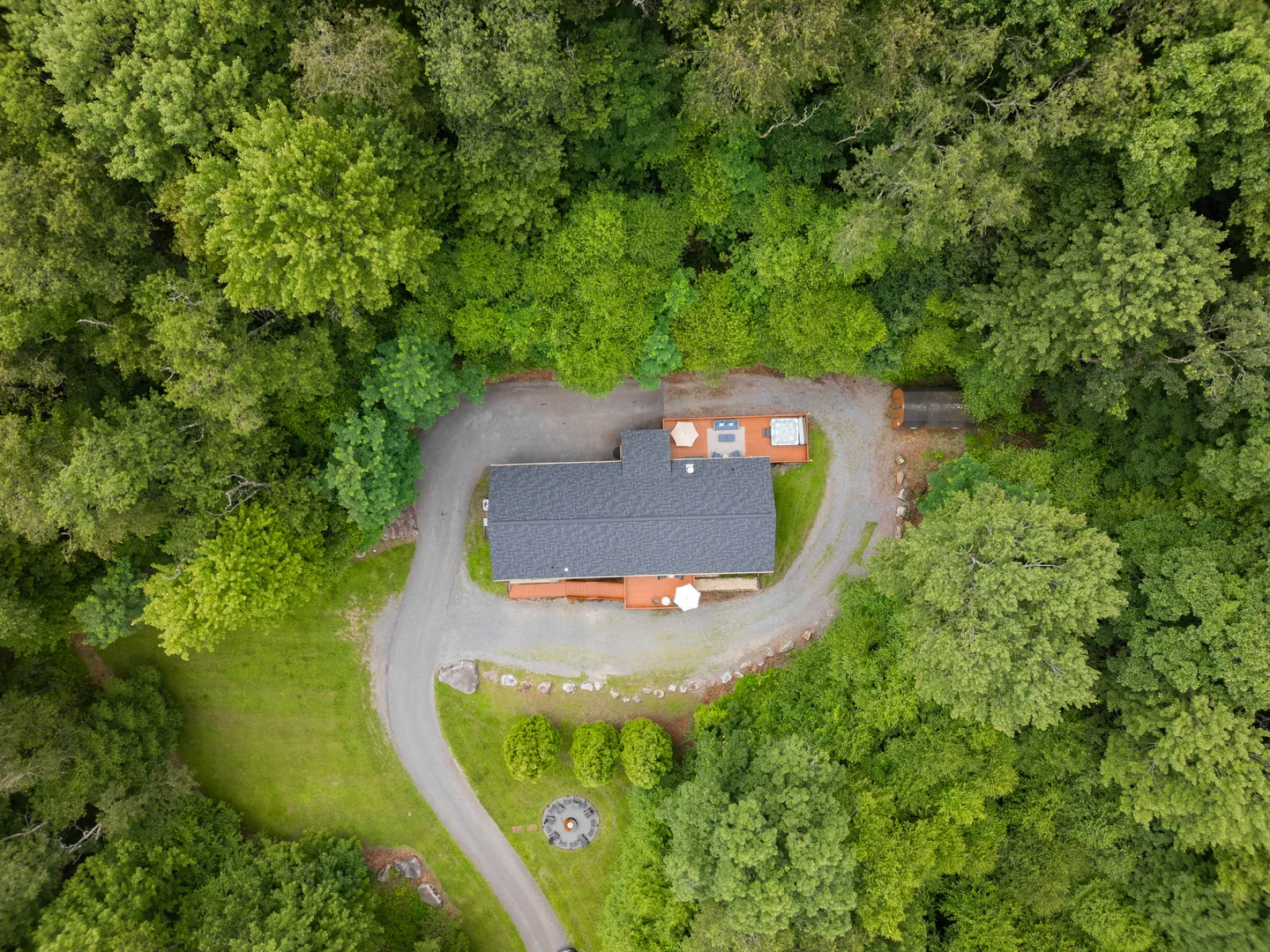 Aerial overhead view of a home surrounded by dense green woods with a gravel driveway, fire pit, and deck.
