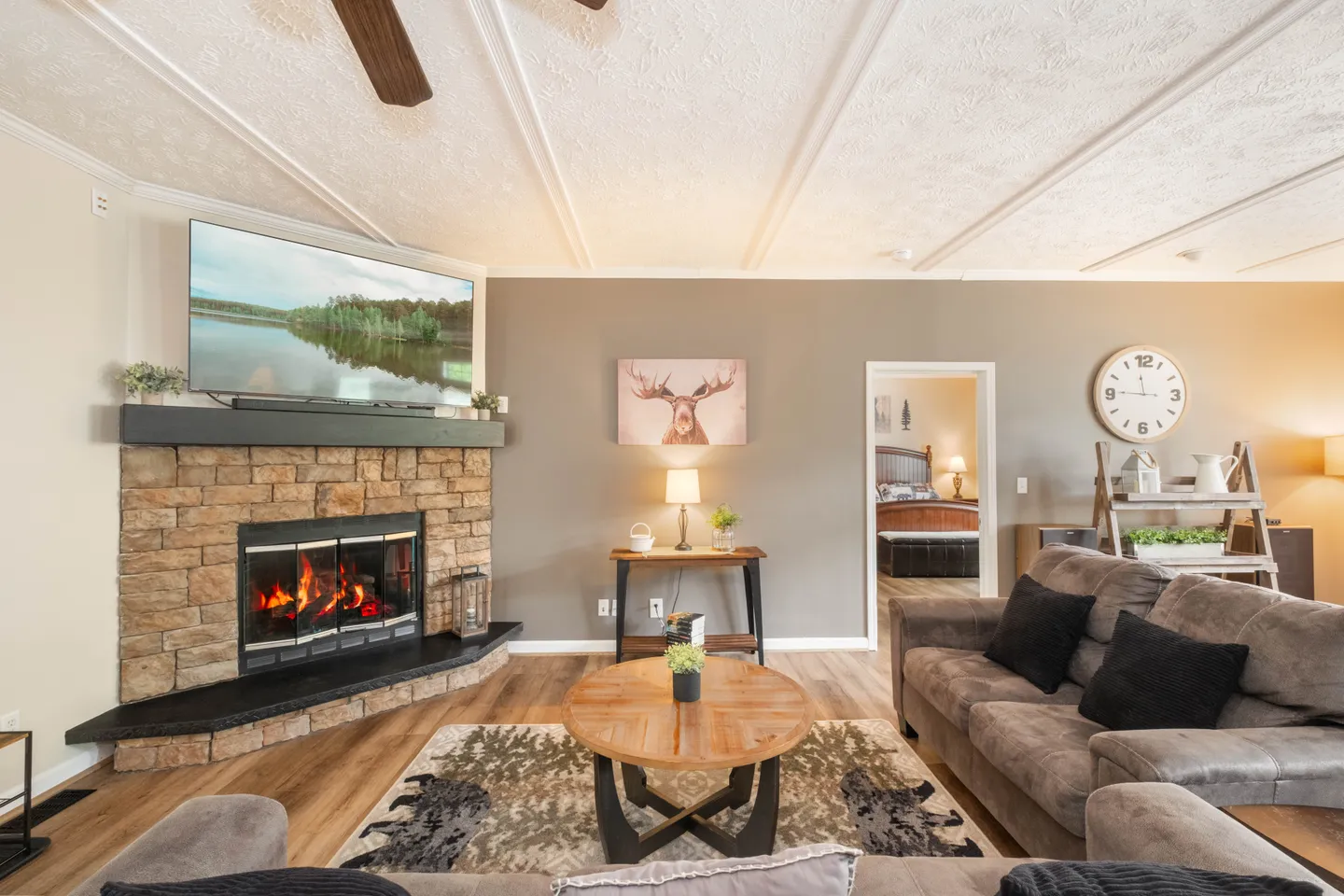 Living room with a stone corner fireplace, mounted TV, plush sofa, circular coffee table, and an open doorway leading to a bedroom.