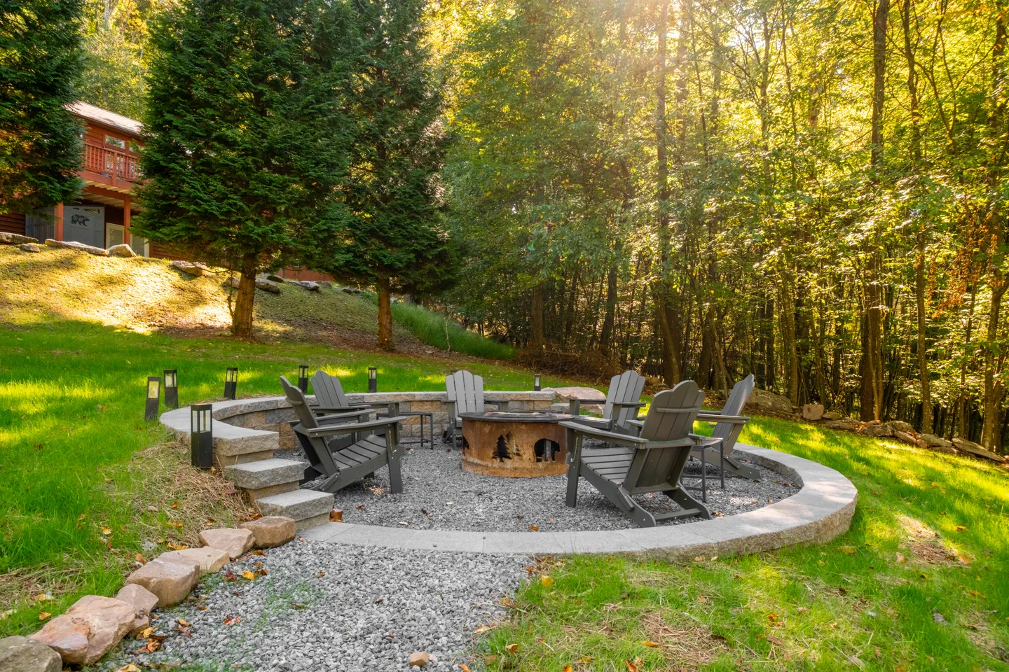 Outdoor gravel fire pit area with Adirondack chairs surrounded by a stone retaining wall and woods.