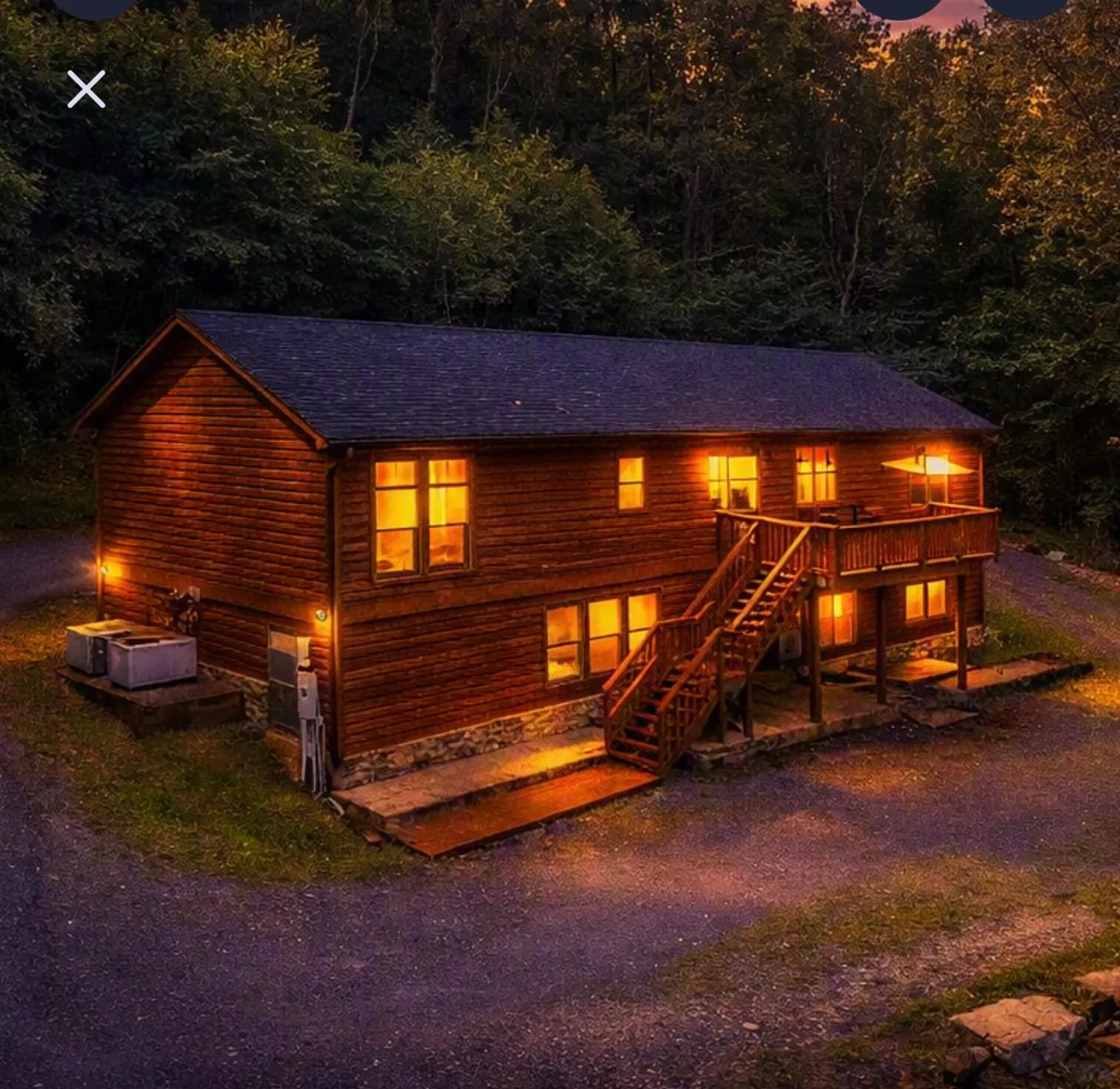 Exterior dusk view of a two-story log cabin surrounded by trees with warm light glowing from the windows and an elevated wooden deck.