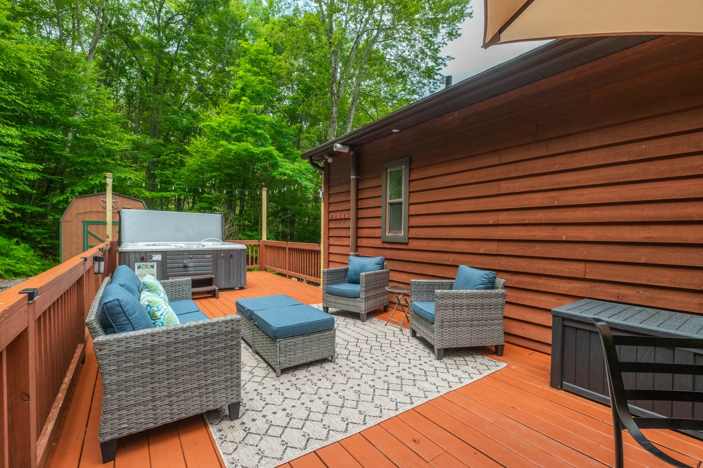 Outdoor wooden deck with wicker seating, an area rug, and a hot tub against a wooded backdrop.