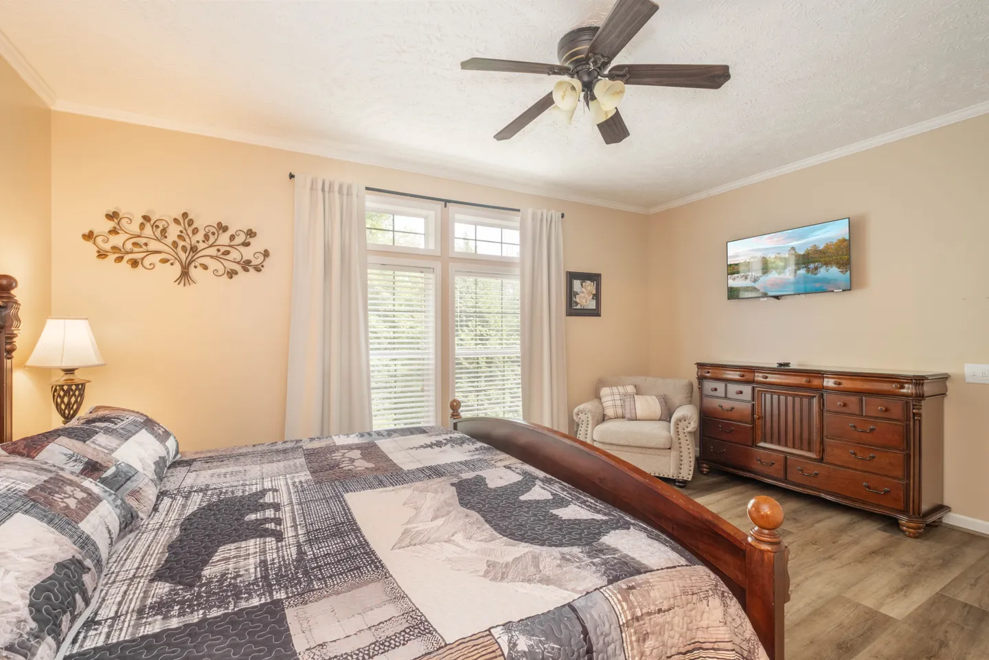 Bedroom featuring a wooden bed with a bear-patterned quilt, a wooden dresser, a mounted TV, and an armchair beside windows.