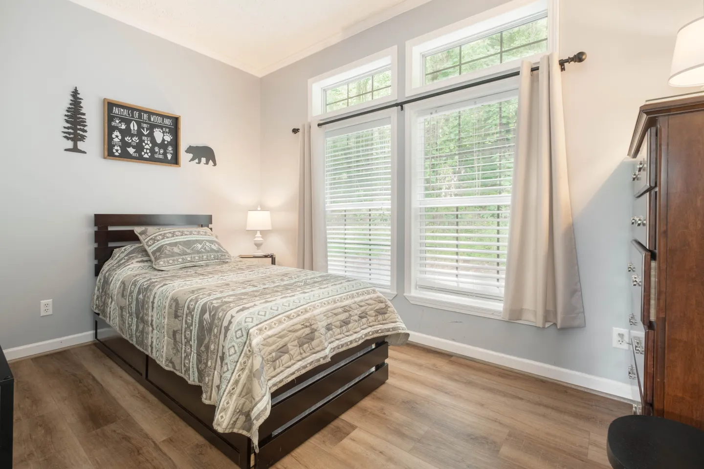 Bedroom featuring a twin bed with a patterned woodland quilt next to large windows with blinds and curtains.