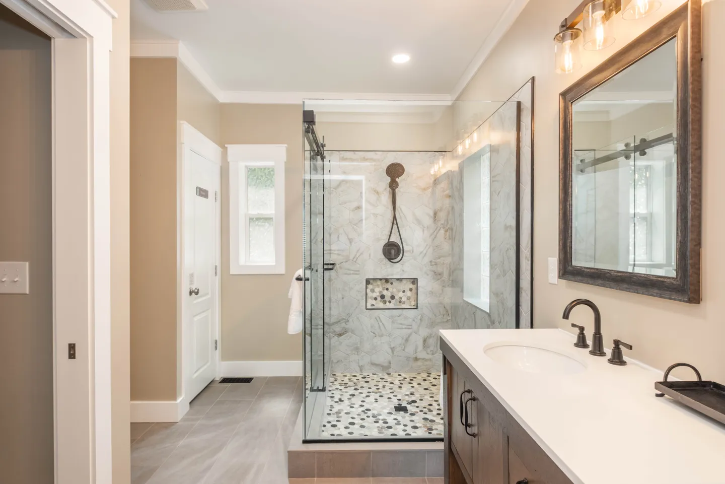 Bathroom with a glass-enclosed walk-in shower, tiled walls, pebble floor, and a vanity with a dark wood-framed mirror.