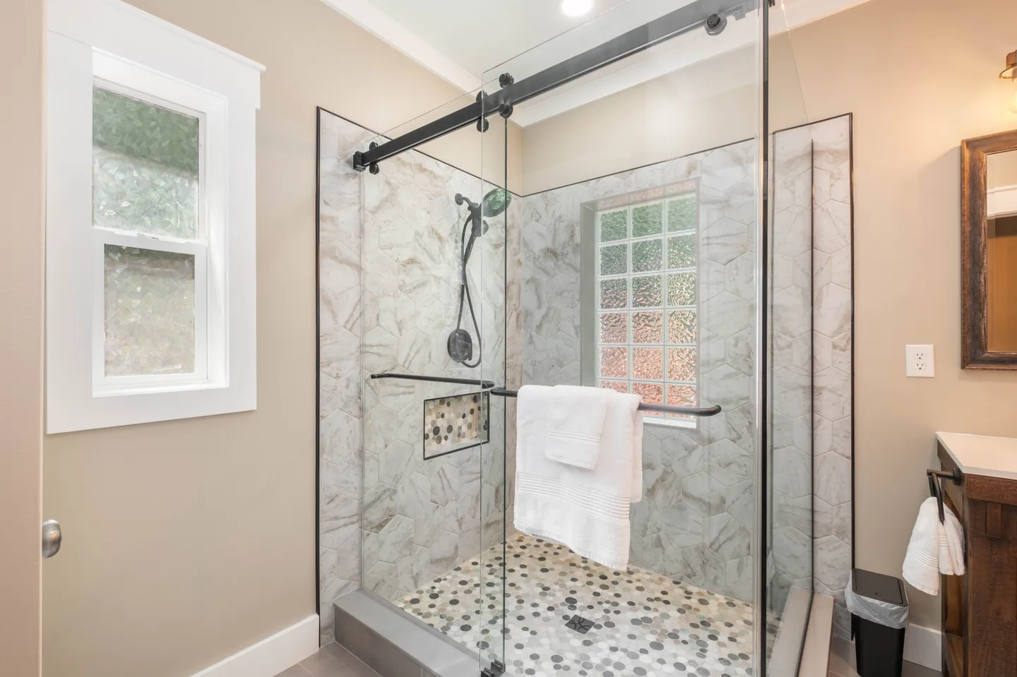 Bathroom walk-in shower with sliding glass doors, dark hardware, pebble-style floor, and a glass-block window.