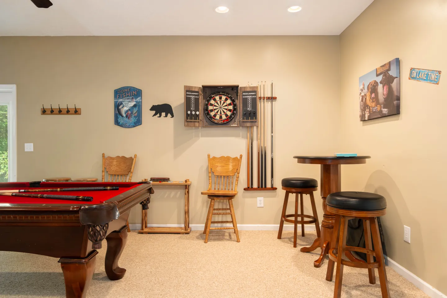 Game room featuring a red felt pool table, dartboard cabinet, cue rack, bar table, and wooden chairs over carpeted flooring.