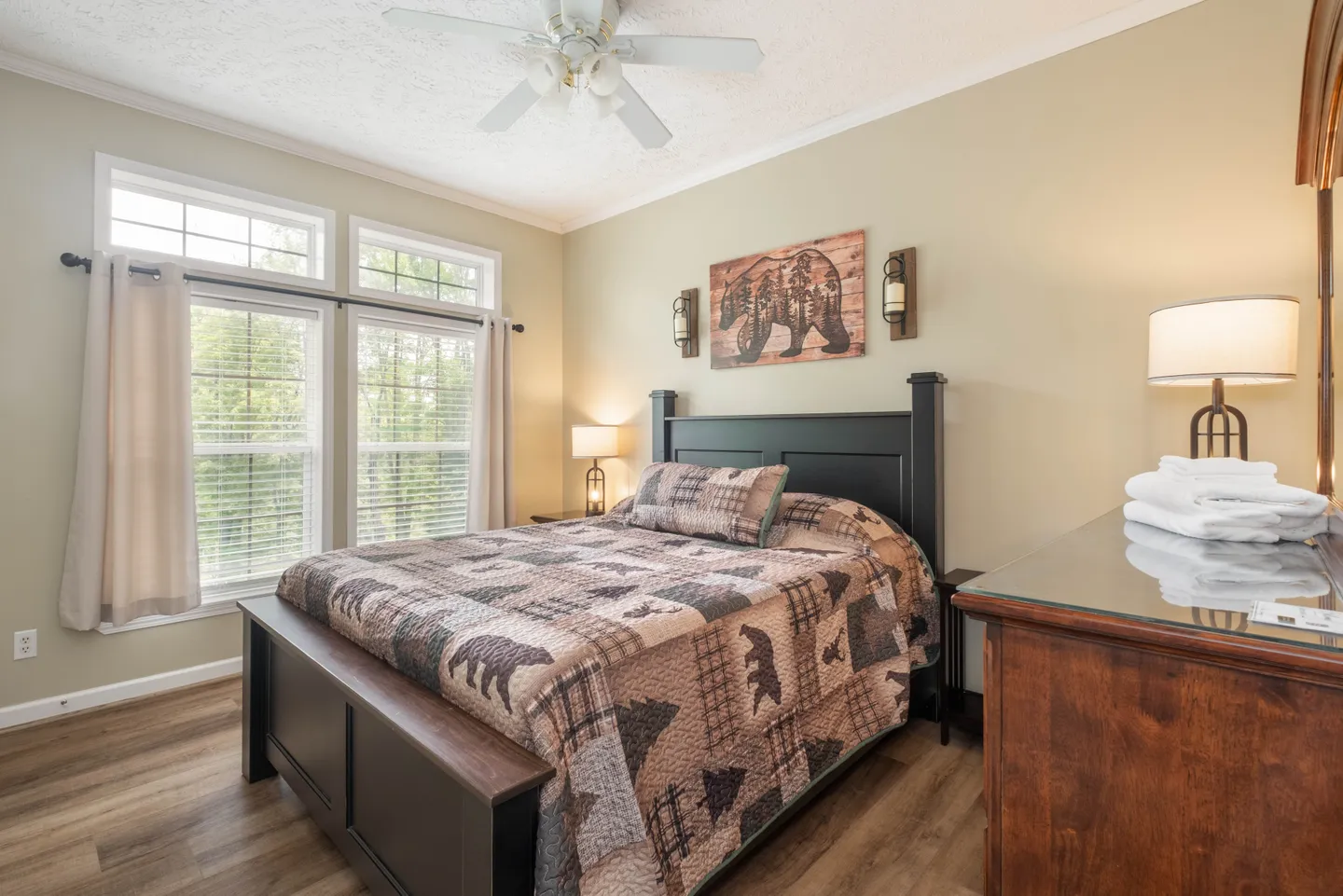 Bedroom with a dark wood bed frame, rustic bear-themed quilt, ceiling fan, and large windows with blinds.
