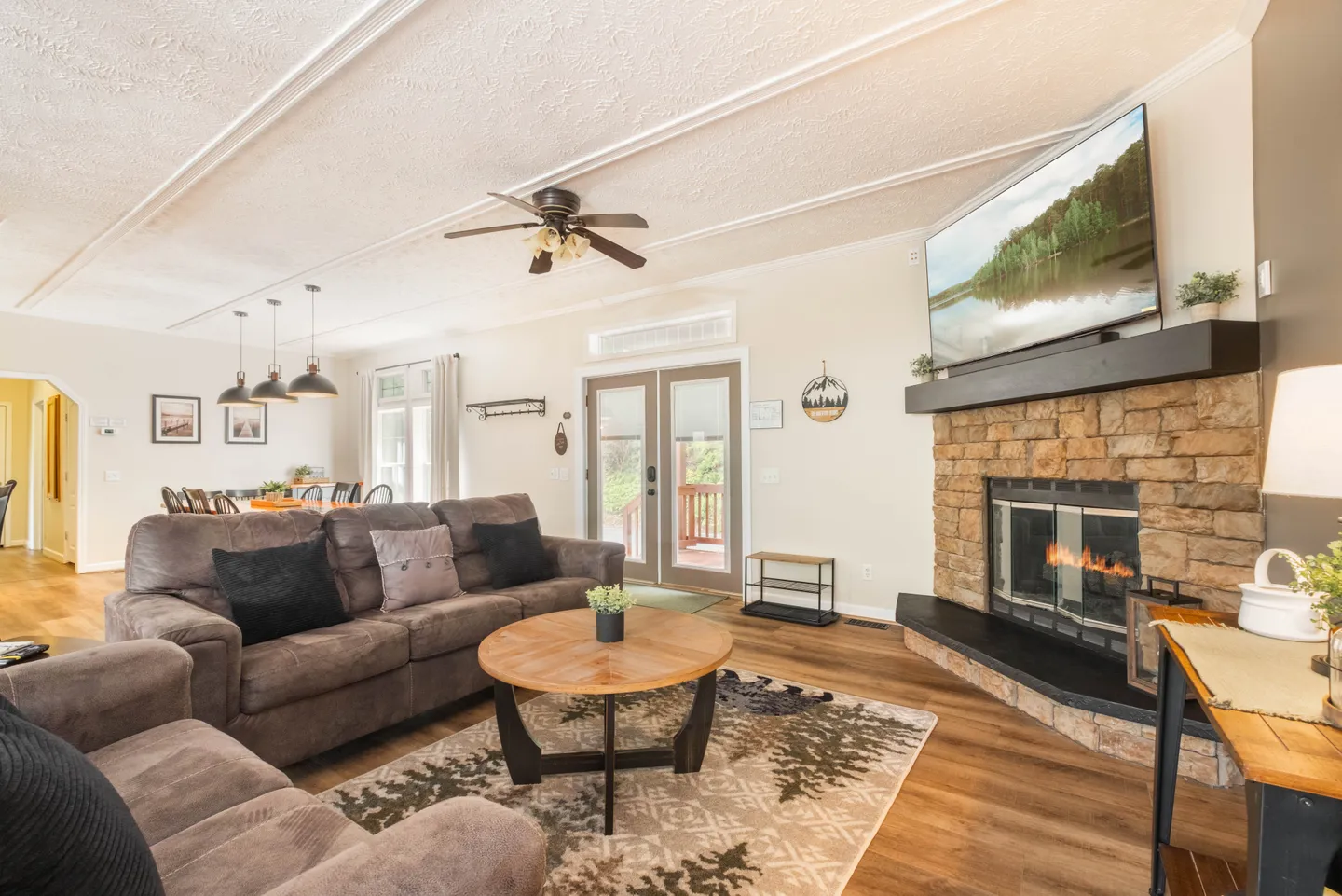 Living room with plush brown sofas, a round wooden coffee table, and a stone-faced fireplace with a television mounted above the mantel.
