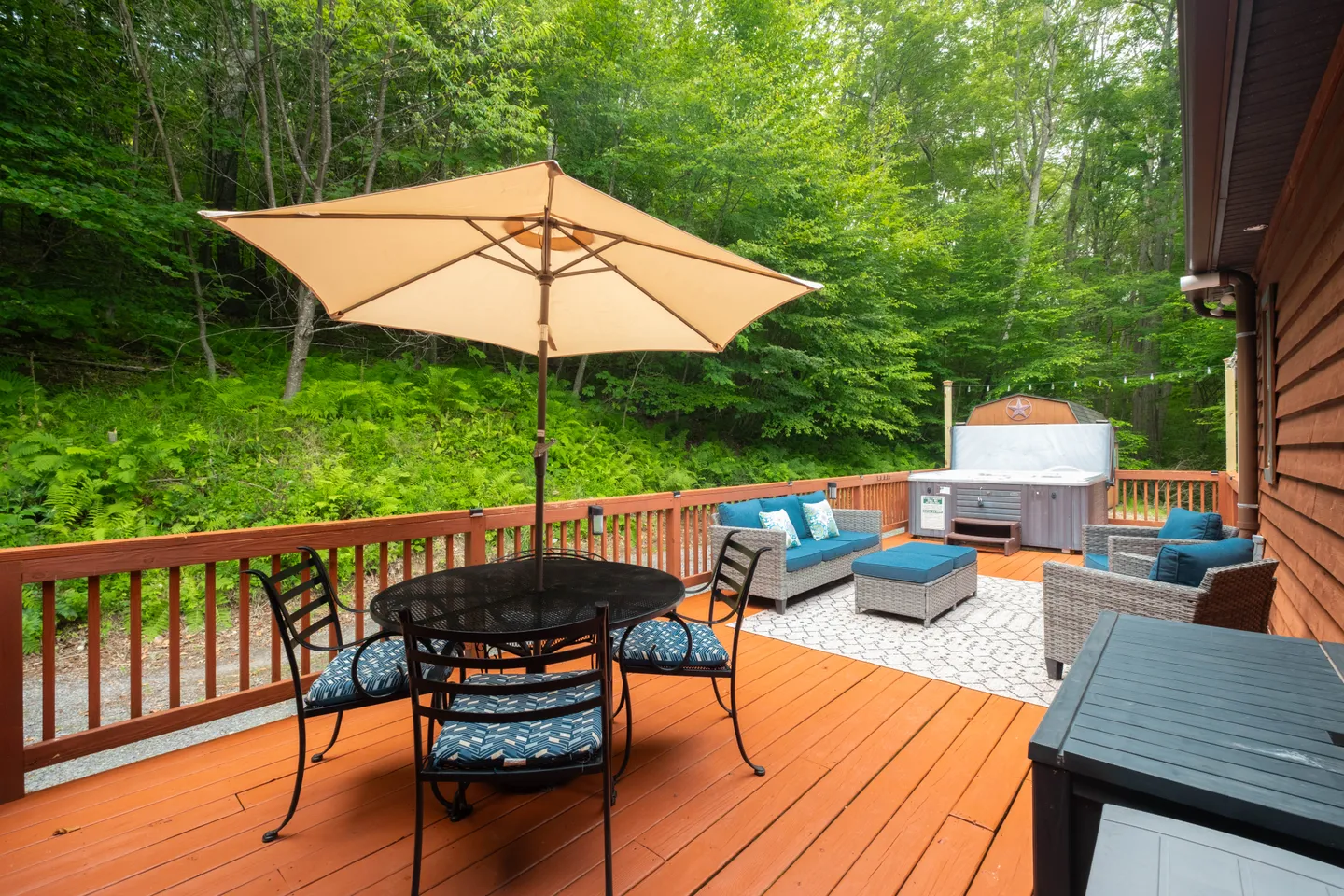Outdoor wooden deck featuring a dining table with an umbrella, lounge seating, and a hot tub surrounded by dense green woods.