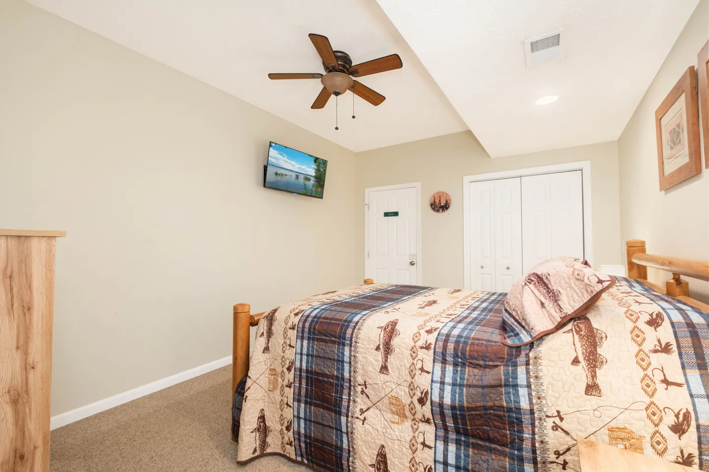 Bedroom featuring a wooden log-style bed with a rustic-patterned quilt, wall-mounted television, ceiling fan, and carpeted floor.