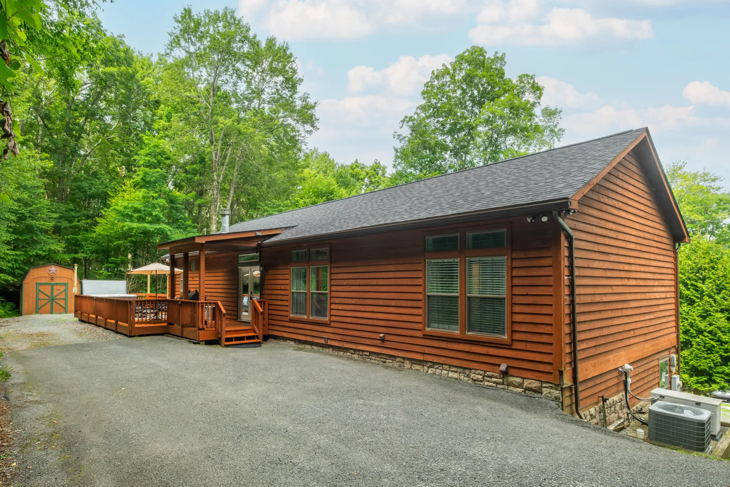 Exterior view of a single-story wood-sided cabin with a back deck, hot tub, and gravel driveway bordered by trees.