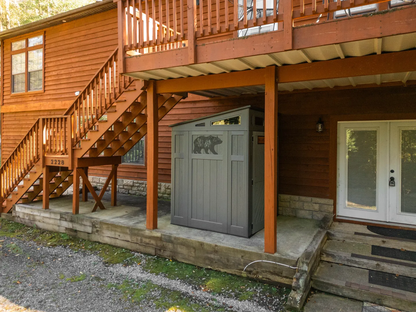 Exterior ground-level view beneath a wooden deck showing an outdoor storage shed with a bear motif, exterior stairs, and an entrance door.