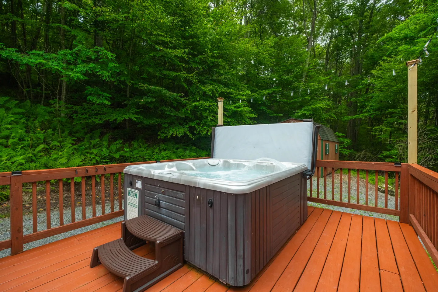 An outdoor hot tub with its cover lifted open on a stained wood deck, bordered by dense green trees.