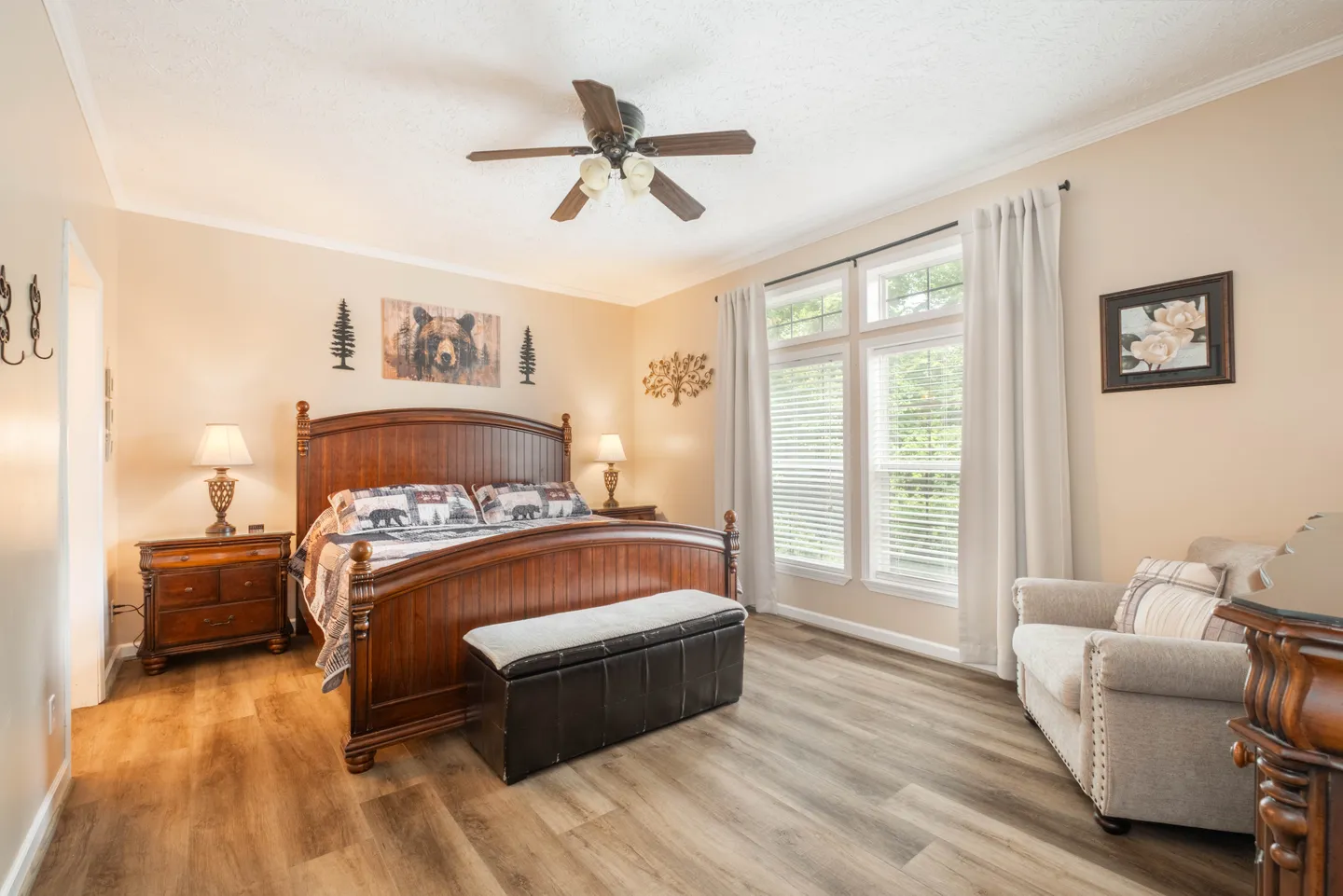Bedroom with a large wooden bed, matching nightstand, storage bench, ceiling fan, and armchair next to two windows with white curtains.