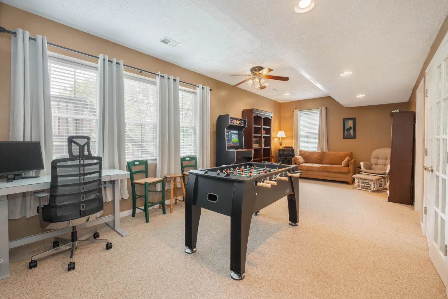 A carpeted recreation and office room featuring a foosball table, an arcade machine, a desk with an office chair, and a seating area with a sofa.