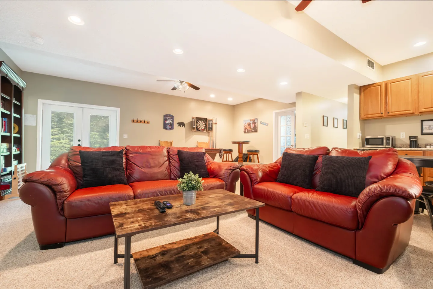 Living area featuring red leather couches, a rustic coffee table, recessed lighting, and a view into a kitchenette and game area with a dartboard.