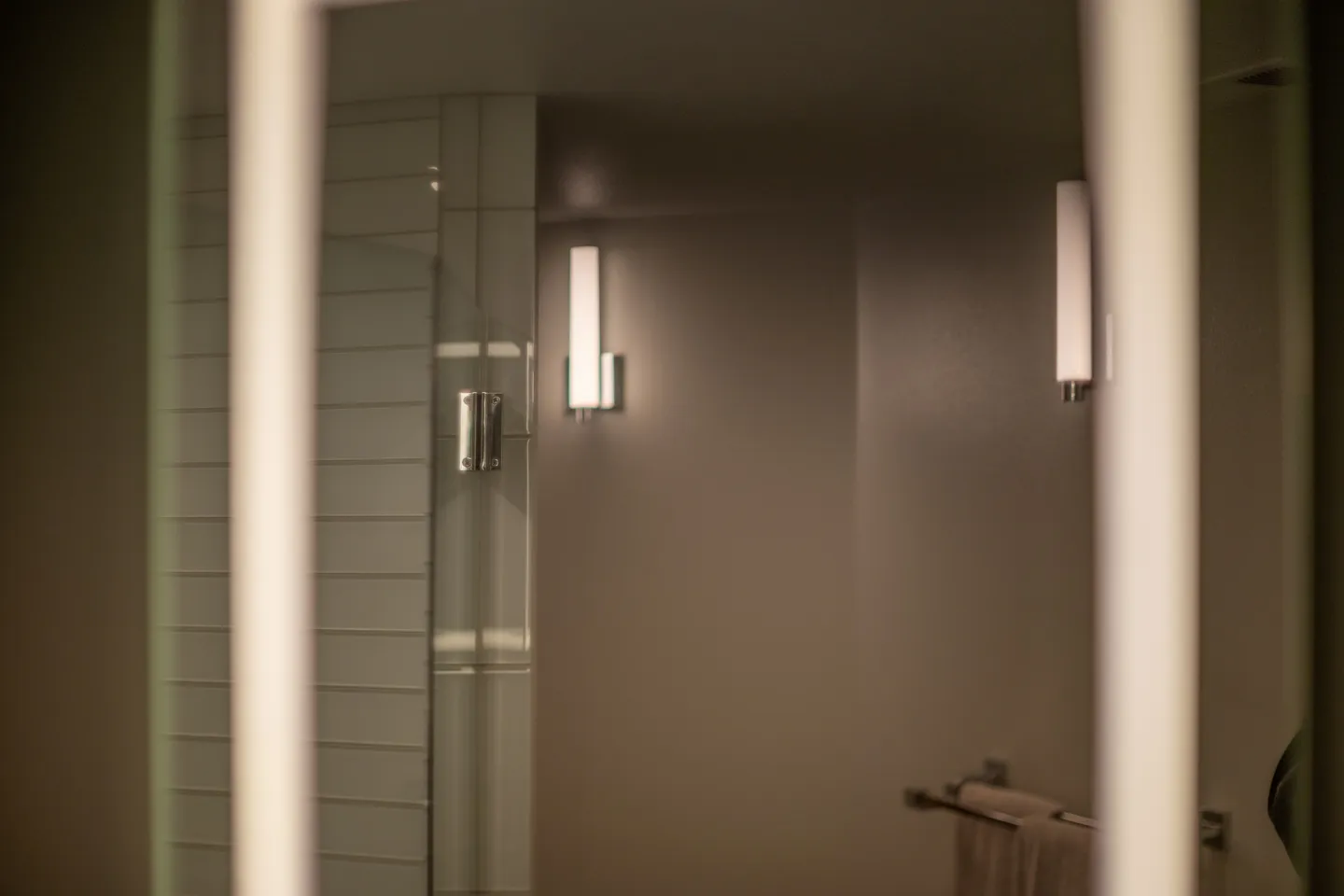 Bathroom mirror reflection showing wall sconce lights and white tile.