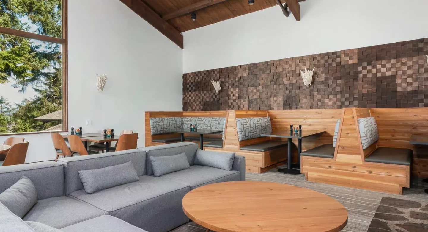 Seating lounge with a grey sectional couch, round wooden coffee table, and built-in wooden dining booths beneath high ceilings.