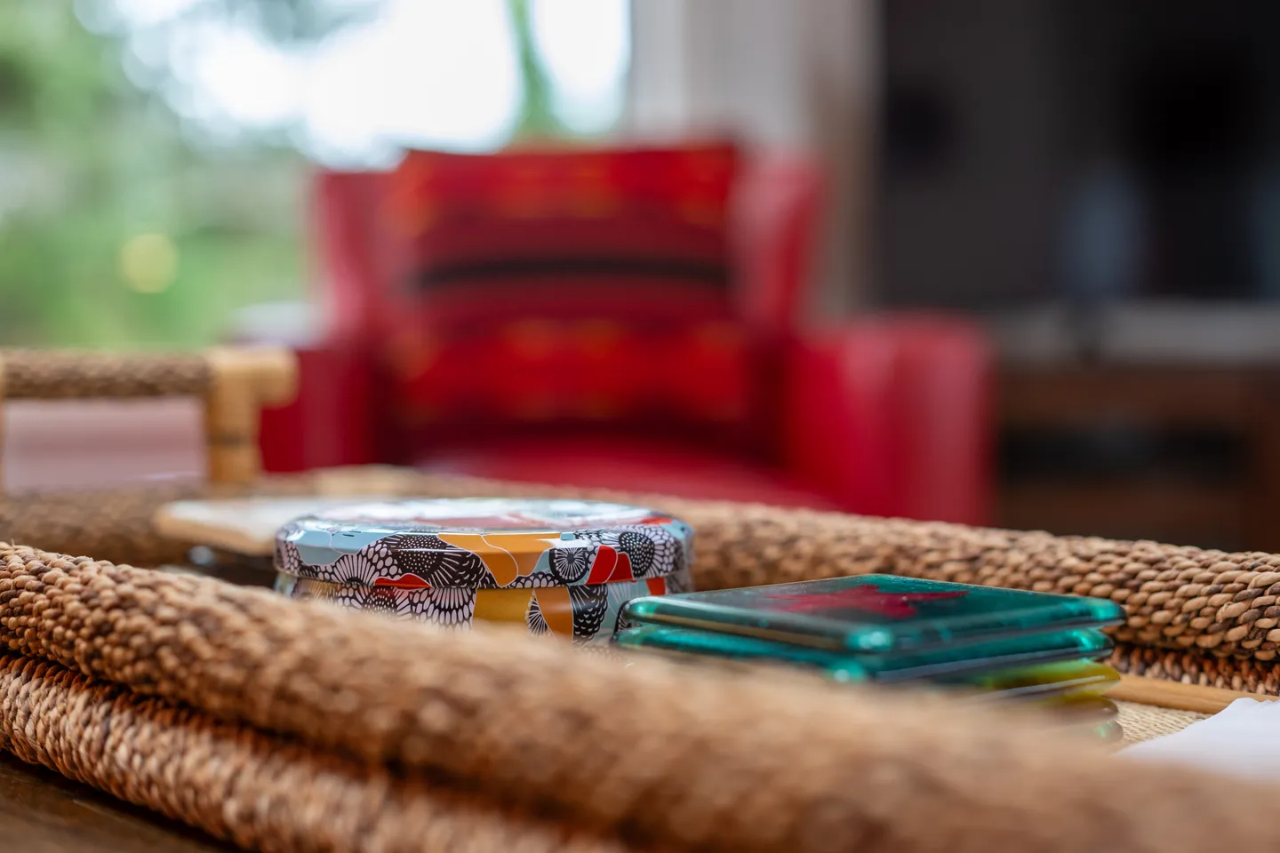 Close-up shot of a woven tray holding coasters and a decorative tin with a blurred red armchair in the background.