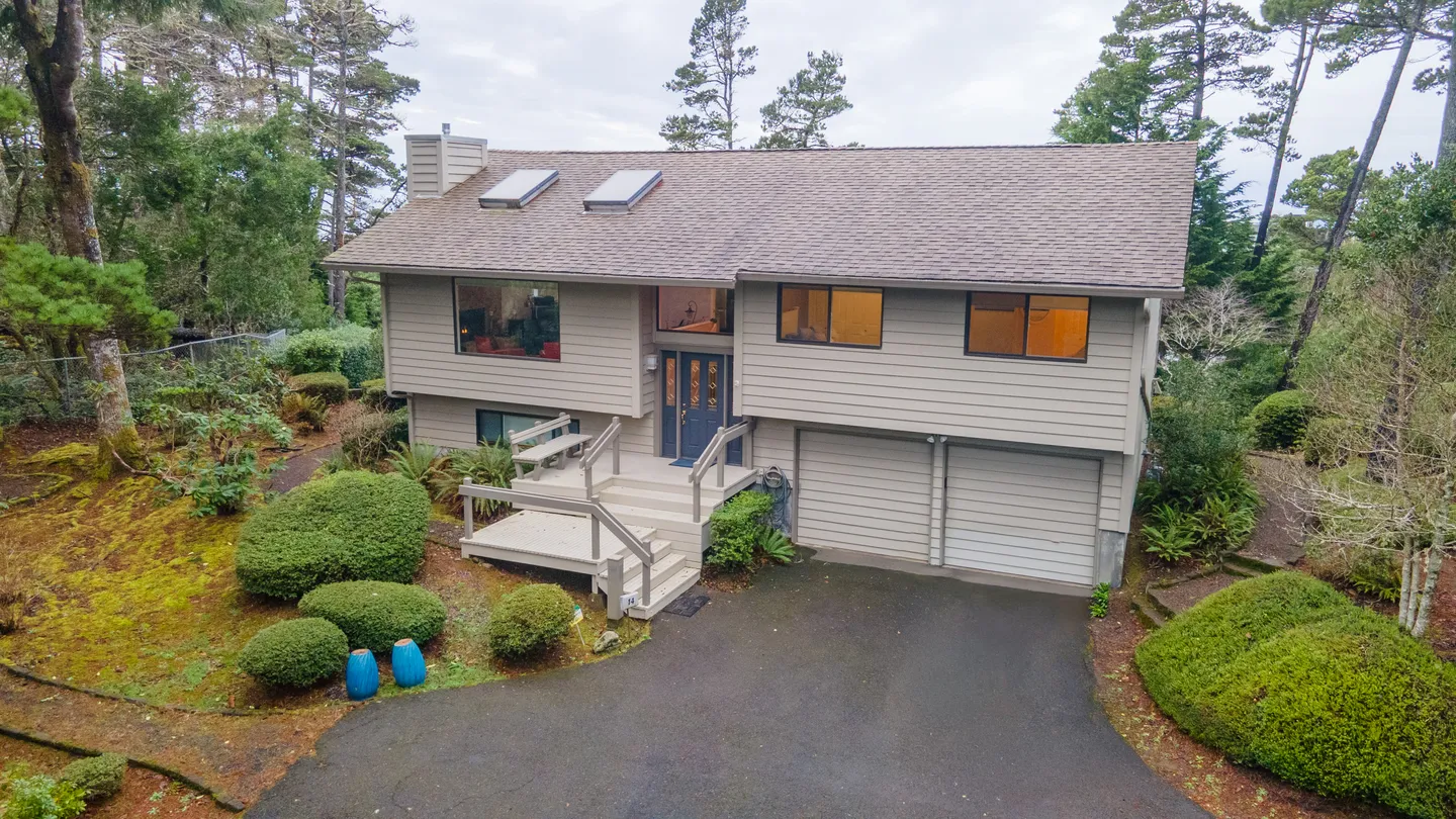 Exterior front view of a split-level home with horizontal siding, an attached two-car garage, wooden entry stairs, and surrounding greenery.