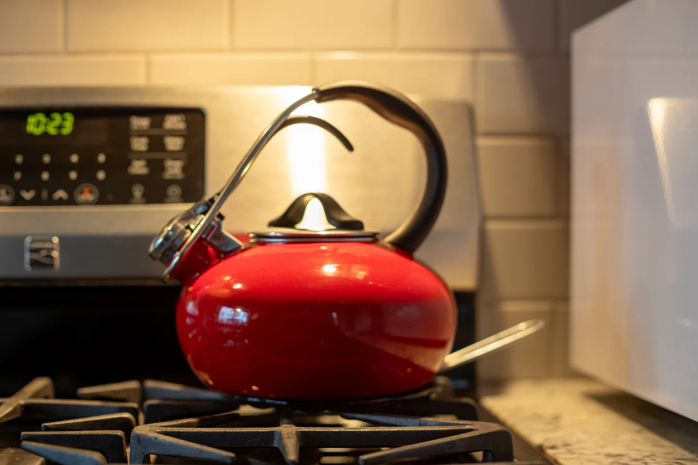 Close-up of a red kettle resting on a gas stove burner in front of white subway tile.