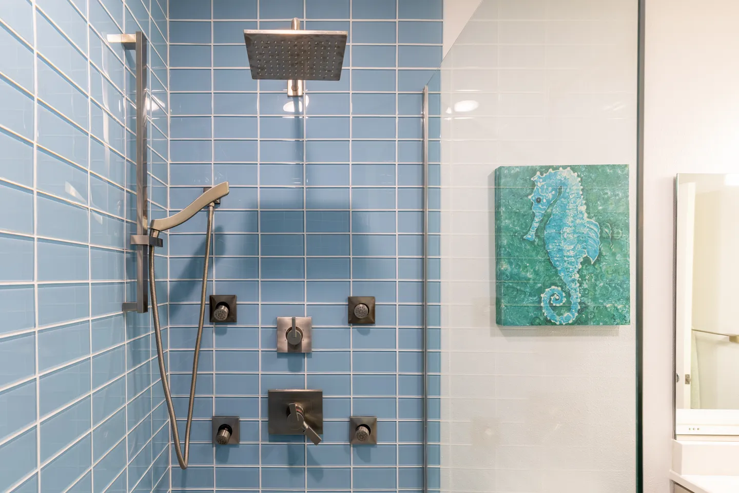 Walk-in shower with light blue vertical subway tile, brushed nickel rainfall showerhead, handheld sprayer, and body jets visible behind a glass enclosure.