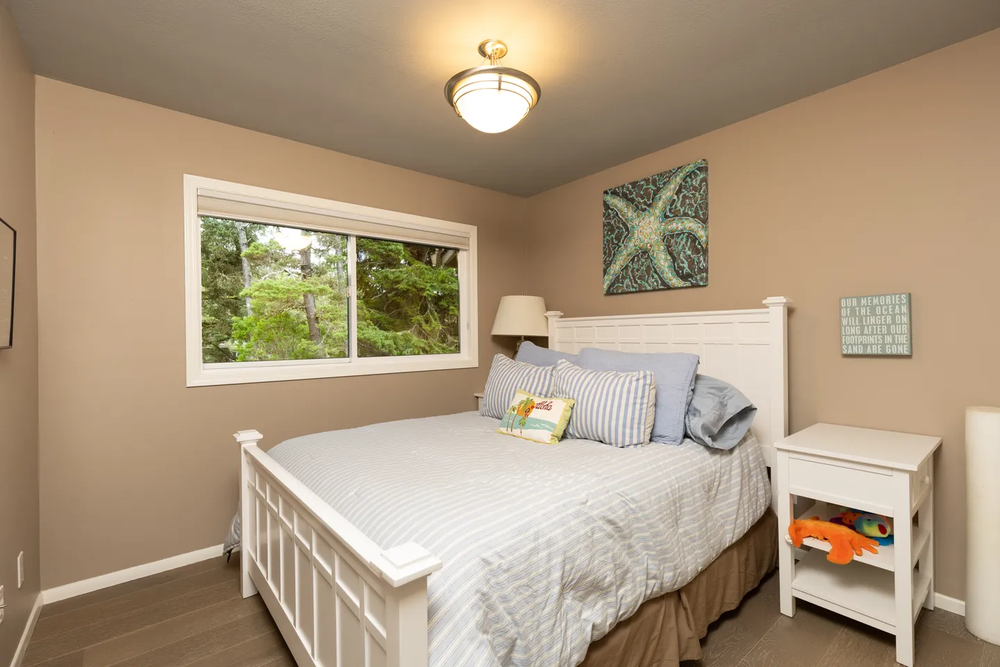 Bedroom with a white panel bed dressed in light blue striped bedding, a white nightstand, and a window overlooking trees.