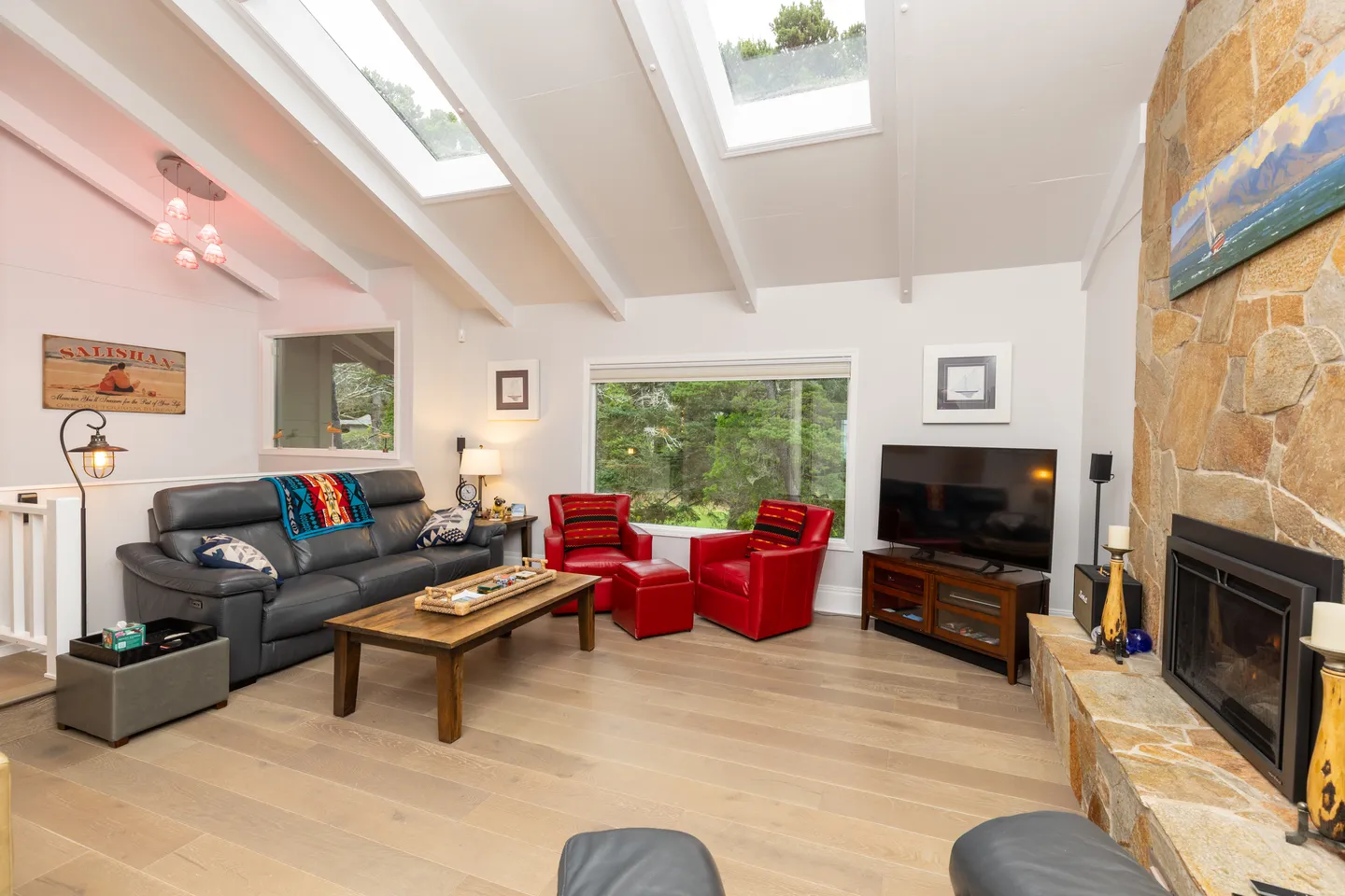 Living room featuring vaulted ceilings with skylights, a stone fireplace, leather sofa, two red armchairs, and a TV.