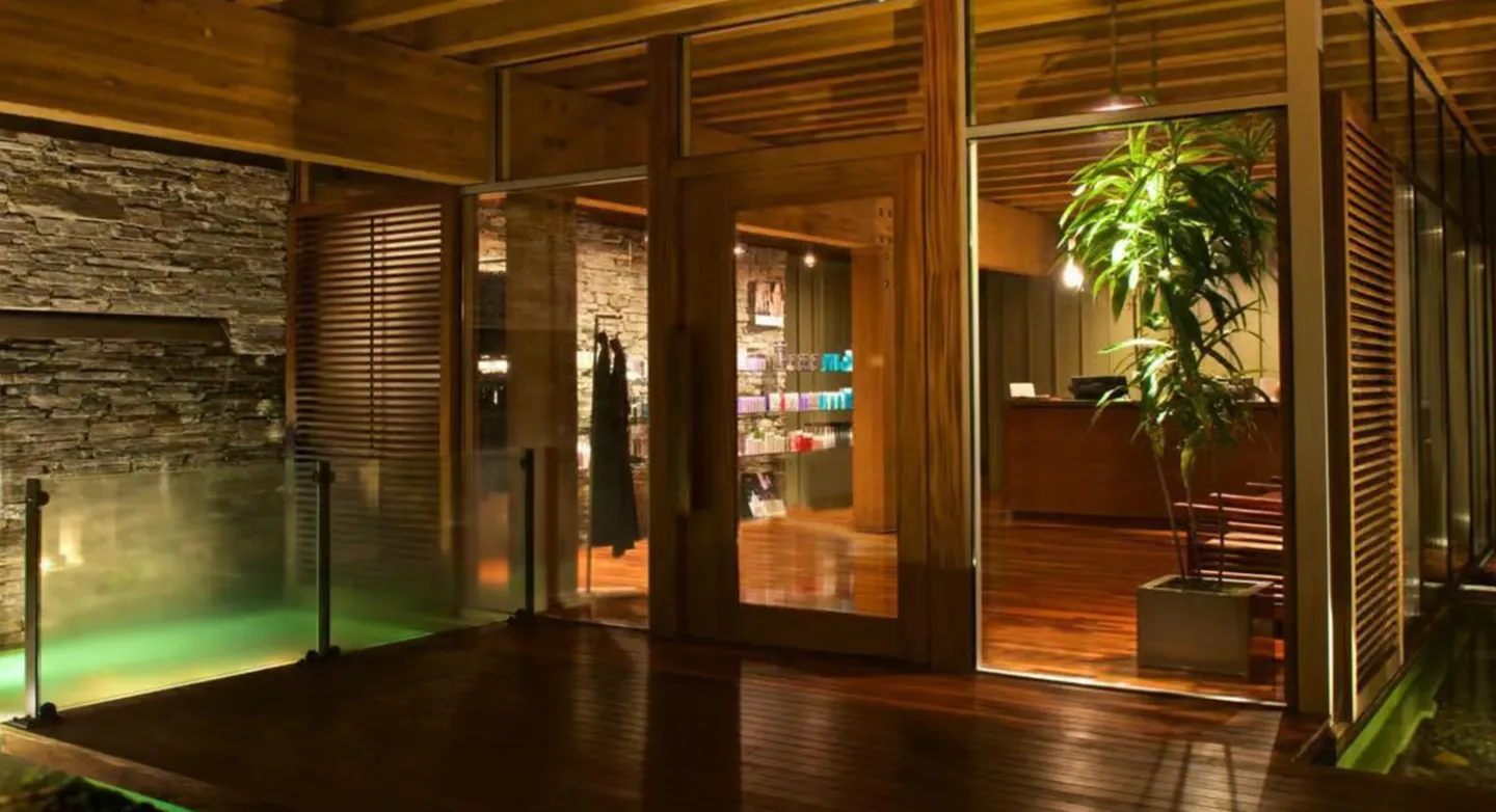 Exterior view through glass walls and door of a warmly lit interior with a reception desk and retail shelves, adjacent to an illuminated water feature.