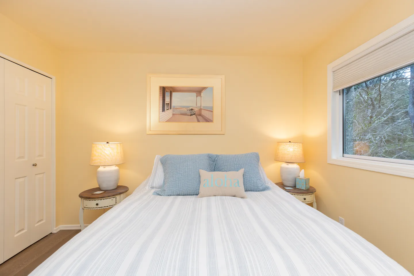 Bedroom with a made bed flanked by nightstands and lamps, warm yellow walls, framed beach artwork, and a window.